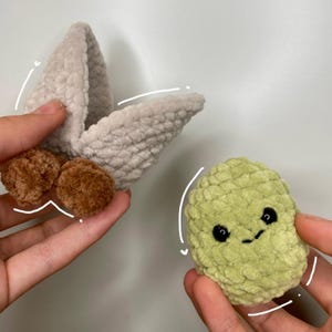 May include: Two handmade crochet items: a light gray, folded shape with brown pom-poms and a small, green, smiling, bean-shaped creature with black eyes. The items are held in hands against a white background.