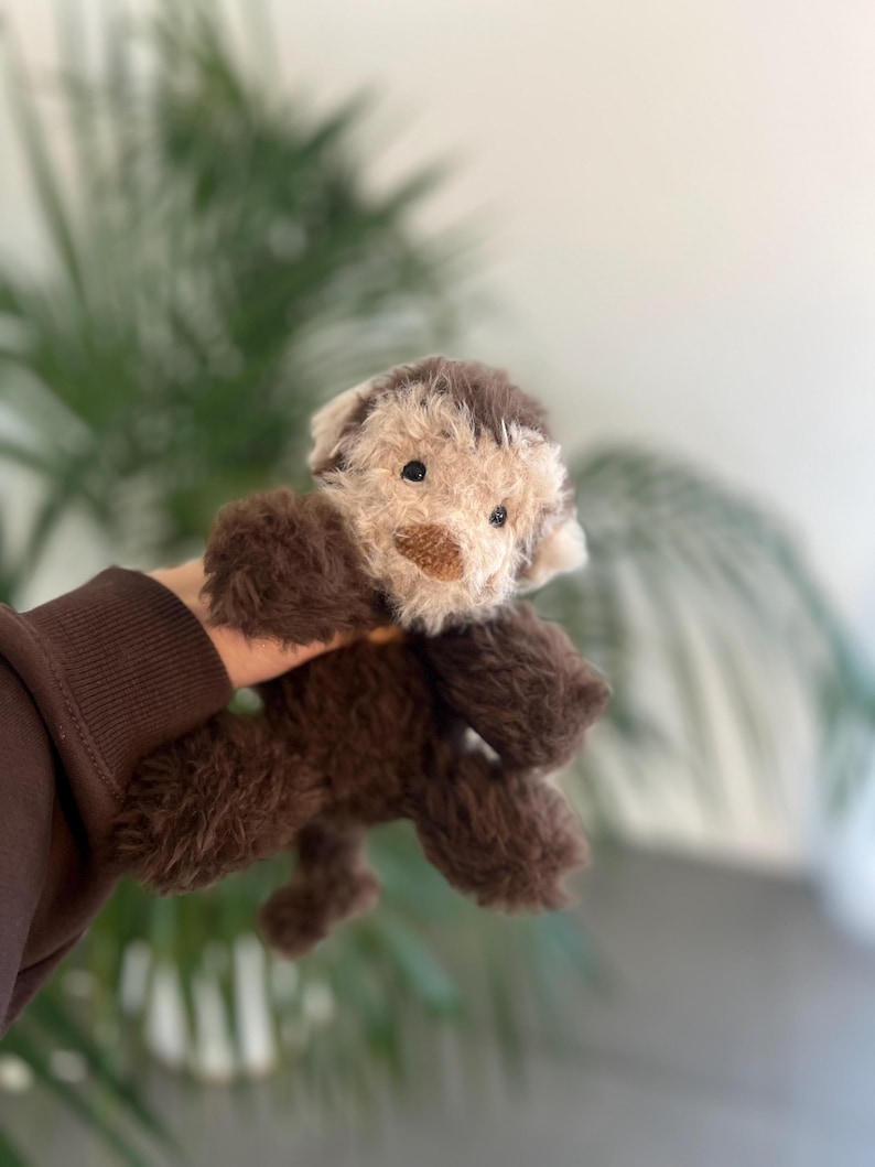 May include: A plush toy dog with brown fur and a tan face. The stuffed animal has a small brown nose and black eyes. The toy is held up against a blurred background of green leaves and a white wall.