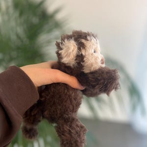 May include: A handmade brown teddy bear with a light beige face and ears. The plush toy has a fluffy texture and small blue eyes. The bear is held in a person's hand, with a blurred green plant background.