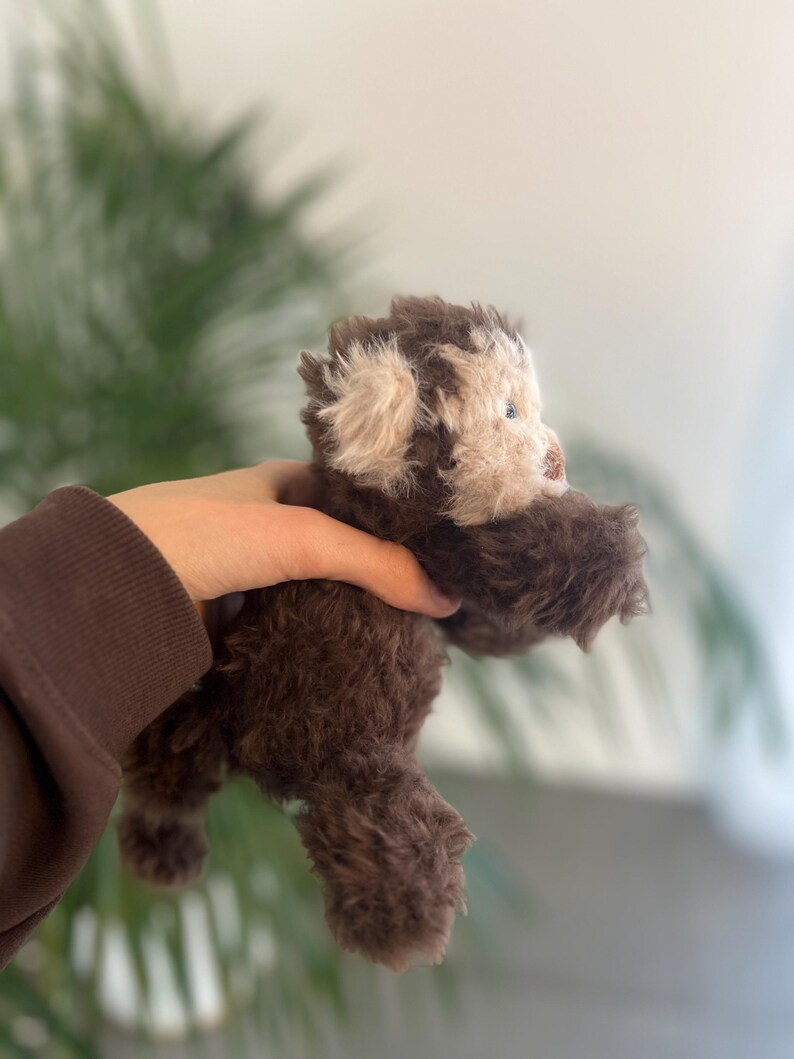 May include: A handmade brown teddy bear with a light beige face and ears. The plush toy has a fluffy texture and small blue eyes. The bear is held in a person's hand, with a blurred green plant background.