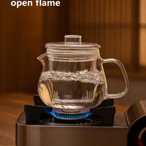 May include: A clear glass teapot with a lid, filled with water, sits on a portable gas stove with a blue flame. The text "Can be heated with an open flame" is visible. The teapot has a handle and spout.