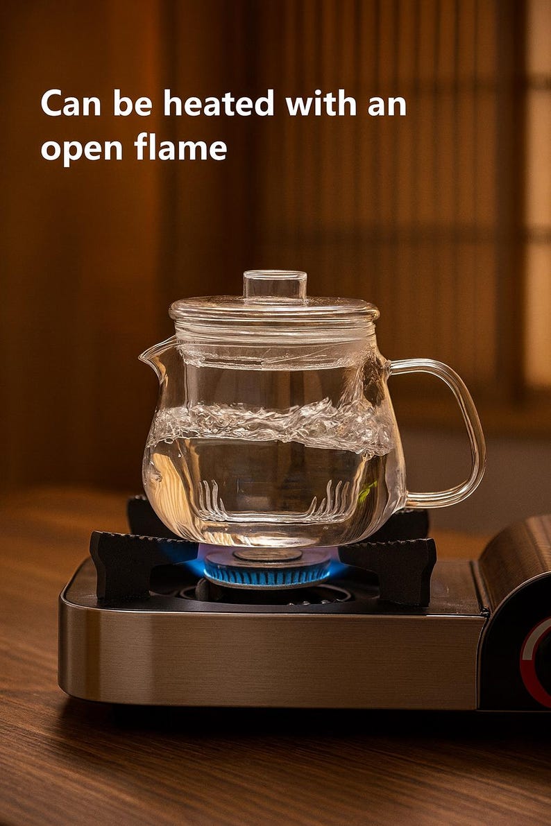May include: A clear glass teapot with a lid, filled with water, sits on a portable gas stove with a blue flame. The text "Can be heated with an open flame" is visible. The teapot has a handle and spout.
