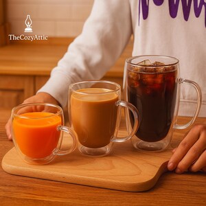 May include: Three clear glass mugs with handles, each filled with a different beverage: orange juice, coffee, and iced coffee. The mugs are on a wooden serving board. The Cozy Attic logo is in the top left corner.
