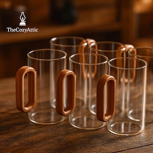 Puede incluir: Mugs de vidrio transparente con asas de madera sobre una superficie de madera. Los mugs son cilíndricos y tienen un diseño sencillo y moderno. Las asas de madera son de color marrón cálido. El logotipo de The Cozy Attic es visible.