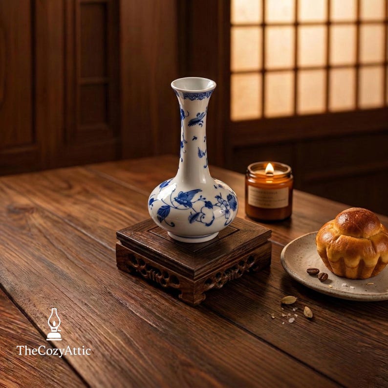 May include: A white and blue porcelain vase with a floral and bird design, displayed on a dark wooden stand. A lit candle and a pastry are also on the wooden table. The Cozy Attic logo is in the bottom left corner.