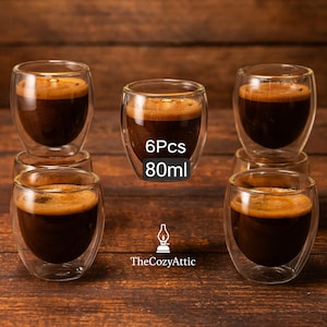 Può includere: Sei tazzine da espresso in vetro trasparente a doppia parete riempite di caffè scuro, appoggiate su una superficie di legno. Ogni tazza contiene 80 ml. Il logo TheCozyAttic è visibile in basso. Le tazze sono progettate per mantenere le bevande calde o fredde.