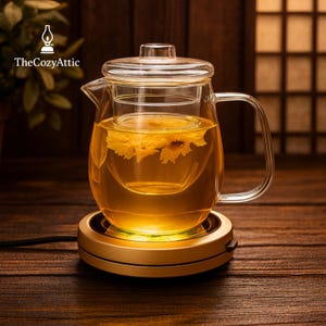 May include: A clear glass teapot with a built-in infuser, filled with golden tea and flowers, sits on a gold-colored warming base. The teapot has a handle and a lid. The Cozy Attic logo is in the upper left corner.