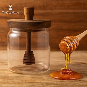 May include: A clear glass honey jar with a wooden lid and dipper. The dipper is dripping honey onto a wooden surface. The jar is next to a wooden honey dipper with honey dripping from it. The Cozy Attic logo is in the top left corner.