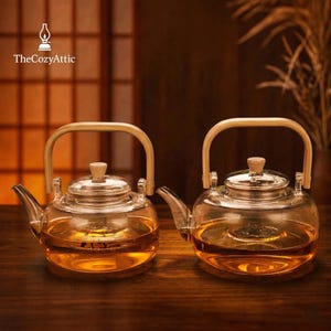 May include: Two clear glass teapots with wooden handles and lids. The teapots are filled with amber-colored tea. The logo "TheCozyAttic" is in the upper left corner. The teapots are on a dark wooden surface.
