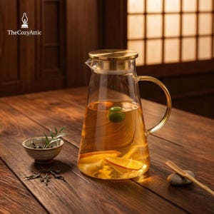 May include: A clear glass pitcher with a gold-colored lid and handle, filled with a light-colored liquid and orange slices. A small ceramic bowl with herbs and loose tea leaves sits on a wooden table. The Cozy Attic logo is in the top left corner.