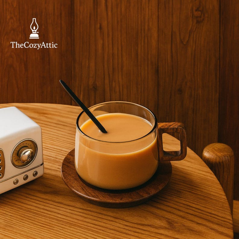 Puede incluir: Una taza de vidrio transparente con asa de madera y cuchara negra, llena de una bebida marr&oacute;n claro, descansa sobre un posavasos de madera. La taza est&aacute; sobre una mesa de madera, junto a una radio blanca. El logotipo de The Cozy Attic es visible.