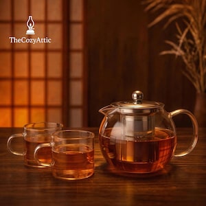 Puede incluir: Una tetera de vidrio transparente con un infusor y tapa de acero inoxidable, llena de té. Dos tazas de vidrio a juego están frente a la tetera. El logotipo "TheCozyAttic" está en la esquina superior izquierda. El juego de té está sobre una superficie de madera.
