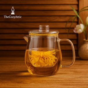 May include: A clear glass teapot with a built-in infuser, filled with golden tea and a yellow flower. The teapot has a handle and a lid. The Cozy Attic logo is in the upper left corner. The background is a wooden surface.