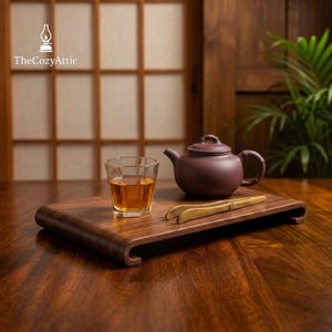 Puede incluir: Una bandeja de té de madera marrón oscuro con un borde curvo sostiene una tetera morada, un vaso de té y un par de pinzas de té doradas. El logotipo de The Cozy Attic está en la esquina superior izquierda. El fondo presenta una pared de madera y una ventana.