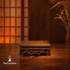 May include: A dark brown wooden display stand with a square top and intricately carved sides. The stand is set on a wooden surface, with a blurred background featuring a window and a vase with dried plants. The Cozy Attic logo is visible.