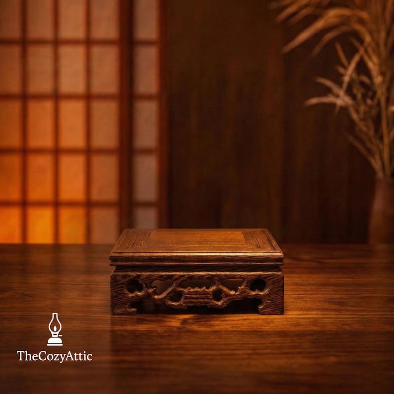 May include: A dark brown wooden display stand with a square top and intricately carved sides. The stand is set on a wooden surface, with a blurred background featuring a window and a vase with dried plants. The Cozy Attic logo is visible.
