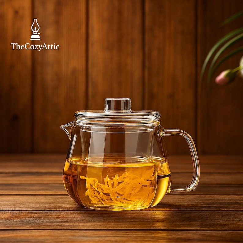 May include: A clear glass teapot with a built-in infuser, filled with golden-colored tea. The teapot has a handle and a lid. The background is a wooden surface. The logo "TheCozyAttic" is in the upper left corner.