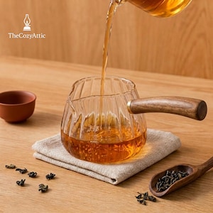 Puede incluir: Una tetera de cristal transparente con estrías verticales y mango de madera, siendo llenada con té ámbar. Una pequeña taza de cerámica marrón, una cuchara de madera con té a granel y hojas de té dispersas sobre un paño de lino claro.