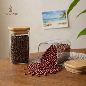 Puede incluir: Dos frascos de vidrio transparente con tapas de bambú. Un frasco está en posición vertical y lleno de granos de pimienta negra. El otro frasco está volcado, derramando frijoles rojos sobre una superficie de madera. Una pequeña foto de una escena de playa está en el fondo.