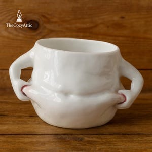 Puede incluir: Una taza de cerámica blanca con un diseño único, con dos asas en forma de brazo y un cuerpo que parece pellizcado. La taza tiene un acabado brillante y está sobre un fondo de madera. El logotipo de The Cozy Attic está en la esquina superior izquierda.