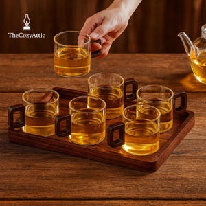 May include: A set of six clear glass mugs with dark brown handles, filled with a golden liquid, presented on a dark wooden tray. The mugs are arranged on a wooden surface, with one mug being held by a hand. The Cozy Attic logo is in the top left corner.