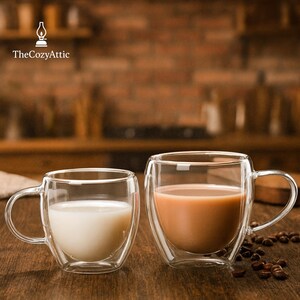 May include: Two clear double-walled glass mugs with curved sides and handles. One mug contains a white liquid, the other a light brown liquid. Coffee beans are scattered near the mugs. The Cozy Attic logo is in the upper left.