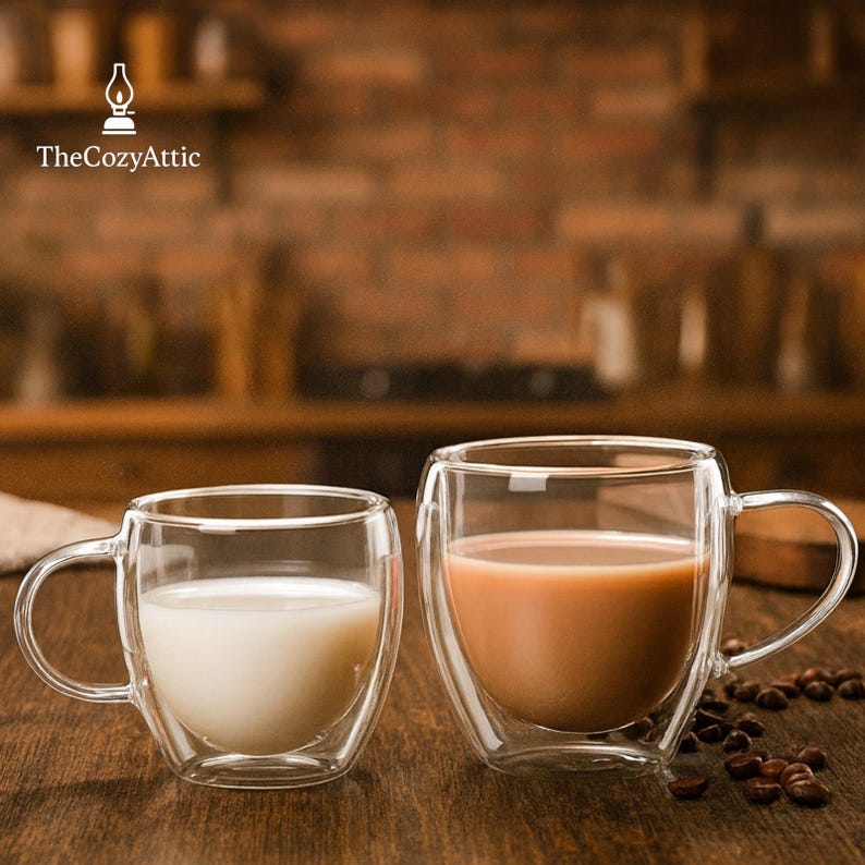May include: Two clear double-walled glass mugs with curved sides and handles. One mug contains a white liquid, the other a light brown liquid. Coffee beans are scattered near the mugs. The Cozy Attic logo is in the upper left.