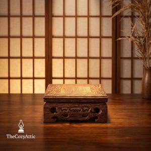 May include: A small, square wooden display stand with carved details. The stand is a warm brown color and sits on a dark wooden surface. The background features a traditional Japanese shoji screen and a vase with dried plants.