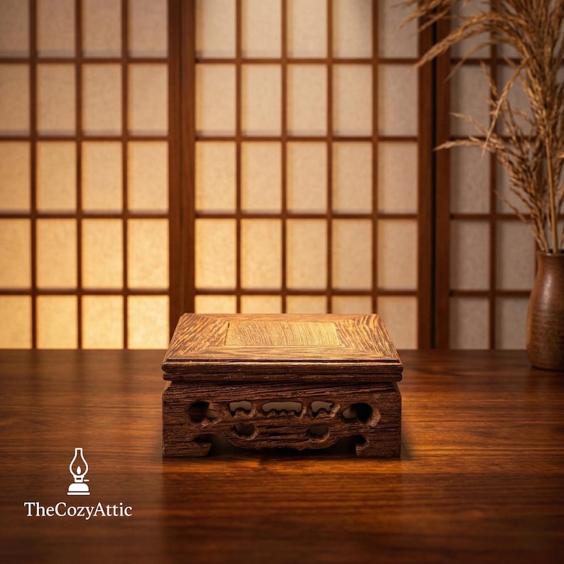 May include: A small, square wooden display stand with carved details. The stand is a warm brown color and sits on a dark wooden surface. The background features a traditional Japanese shoji screen and a vase with dried plants.