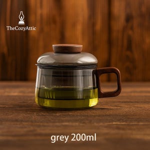 Puede incluir: Taza de cristal para infusi&oacute;n de t&eacute; con asa y tapa de madera. La taza contiene t&eacute; verde y tiene un borde gris. El texto "TheCozyAttic" est&aacute; en la esquina superior izquierda y "grey 200ml" en la parte inferior.
