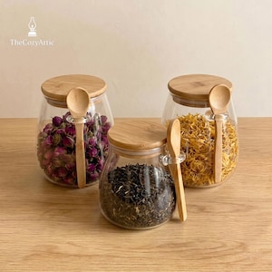 May include: Three glass jars with wooden lids and spoons. The jars contain dried herbs and flowers in pink, yellow, and black. The jars are on a wooden surface. The text "The CozyAttic" is in the upper left corner.