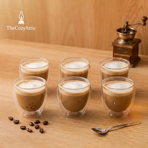 Könnte beinhalten: Sechs klare, doppelwandige Kaffeetassen aus Glas, gefüllt mit Kaffee und mit weißem Schaum bedeckt. Kaffeebohnen und ein kleiner Löffel liegen auf der Holzoberfläche. Eine Vintage-Kaffeemühle befindet sich im Hintergrund. Das Cozy Attic-Logo befindet sich oben links.