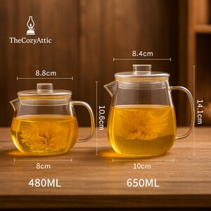 May include: Two glass teapots with lids, filled with yellow liquid and a flower. The smaller teapot is 8 cm wide and holds 480ml. The larger teapot is 10 cm wide and holds 650ml. The Cozy Attic logo is in the upper left corner.