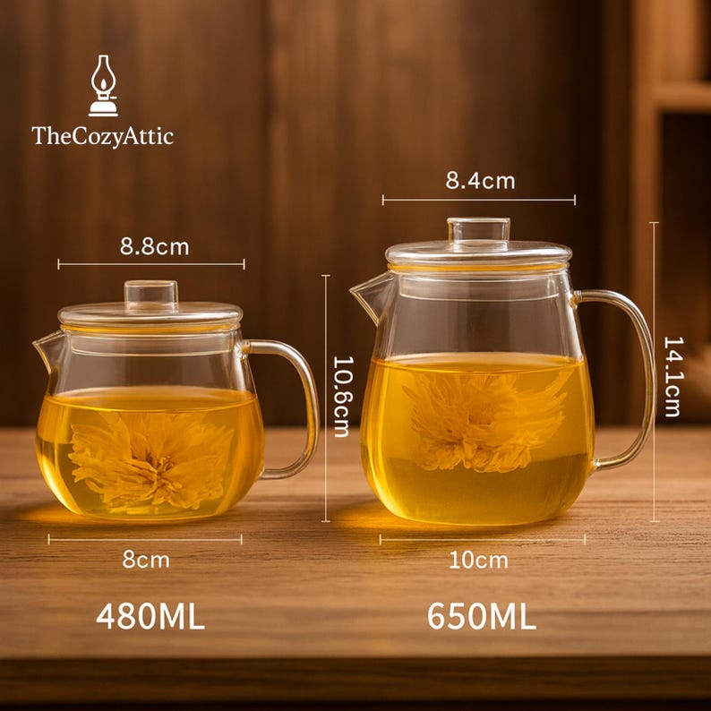 May include: Two glass teapots with lids, filled with yellow liquid and a flower. The smaller teapot is 8 cm wide and holds 480ml. The larger teapot is 10 cm wide and holds 650ml. The Cozy Attic logo is in the upper left corner.