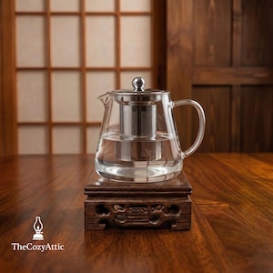 May include: A clear glass teapot with a stainless steel infuser and lid, filled with water. The teapot sits on a dark brown wooden stand with carved details. The Cozy Attic logo is in the bottom left corner.