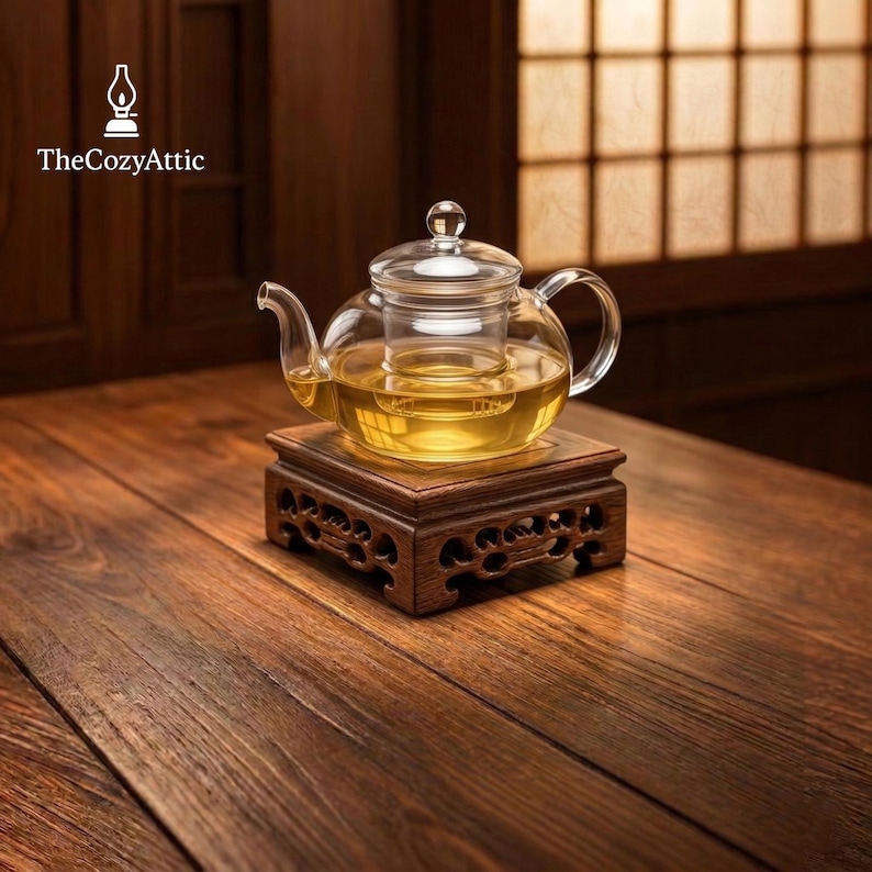May include: A clear glass teapot with a glass infuser, filled with tea, sits on a carved wooden stand. The teapot has a rounded shape with a handle and spout. The stand is dark brown and has intricate carvings. The Cozy Attic logo is in the upper left corner.