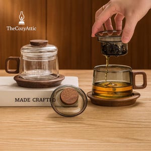 Könnte beinhalten: Klarer Teeglasbecher mit Holzgriff und Untersetzer. Eine Hand gießt Tee aus einem Metall-Infuser in die Tasse. Die Tasse ist mit Tee gefüllt. Ein weiterer Teeglasbecher mit Deckel und Holzuntersetzer ist ebenfalls zu sehen. Der Text "MADE CRAFTED" ist vorhanden.