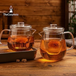 Puede incluir: Dos teteras de vidrio transparente con infusores, llenas de té color ámbar y una flor. Cada tetera tiene un asa y una tapa. Las teteras están sobre una superficie de madera. El logotipo de The Cozy Attic está en la esquina superior izquierda.
