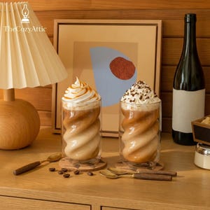 Könnte beinhalten: Zwei klare doppelwandige Gläser mit gedrehtem Spiraldesign, gefüllt mit einem Kaffeegetränk. Eines ist mit Schlagsahne und Karamellsauce, das andere mit Schlagsahne und Schokoladenraspeln garniert, platziert auf Untersetzern aus Kork.