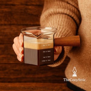 May include: A clear glass measuring cup with a wooden handle, filled with coffee. The cup has measurement markings in ounces and milliliters. The coffee is layered with a dark brown base and a lighter tan foam. The Cozy Attic logo is in the bottom right corner.