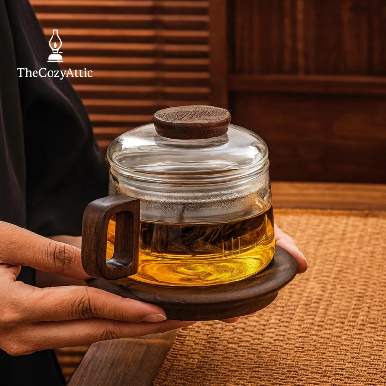 Puede incluir: Una taza de t&eacute; de vidrio transparente con asa, tapa y base de madera. La taza est&aacute; llena de t&eacute; y est&aacute; sobre un posavasos de madera. El logotipo de The Cozy Attic est&aacute; en la esquina superior izquierda.