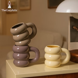 Puede incluir: Tres tazas de cerámica con un diseño apilado único. Una taza es de color amarillo claro, otra morada y la tercera marrón claro. Cada taza tiene un asa redondeada y un cuerpo cilíndrico con secciones redondeadas apiladas. El logotipo "TheCozyAttic" está en la esquina superior izquierda.