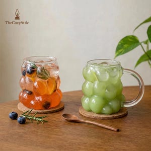 Puede incluir: Dos tazas de vidrio transparente con un diseño de burbujas único, cada una llena de una bebida refrescante. Una taza contiene una bebida naranja con frutas y hierbas, la otra una bebida verde. Ambas están sobre posavasos de corcho, con una cuchara de madera cerca.