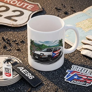 May include: White ceramic mug featuring a car with a Puerto Rican flag design, palm trees, and a mountain landscape. Includes a coaster, keychain, and sticker with car-related graphics. A Route 22 sign and a map are in the background.