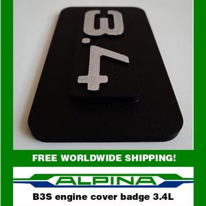 May include: Black engine cover badge with the number "3.4" in silver. The badge is rectangular with a smaller, raised section. The text "ALPINA" and "B3S engine cover badge 3.4L" are at the bottom.