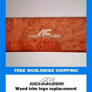 May include: A rectangular wood trim piece with a burl wood grain pattern. The trim features the "AC Schnitzer" logo in white. The text "Wood trim logo replacement" is below the logo. The background is white with a blue border.