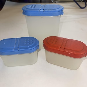 May include: Three plastic spice containers with translucent white bodies and colored lids. One container has a blue lid, and another has a red lid. The third container has a blue lid. Each lid has a sliding mechanism and small holes for dispensing.