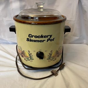 Puede incluir: Una olla de cocción lenta eléctrica de color crema con borde marrón y tapa de cristal transparente. El frente muestra el texto Crockery Simmer Pot, una perilla negra con ajustes Off, Low, High y un gráfico colorido de vegetales.