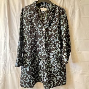 Puede incluir: Un vestido abotonado de manga larga, con un fondo oscuro y un estampado floral repetido en tonos azules y verdes. El vestido tiene cuello y cintura entallada. La tela parece ser de algodón o lino ligero.