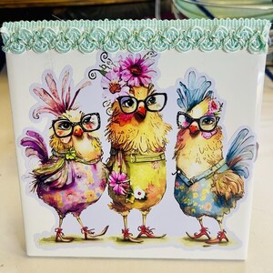 Op de afbeelding: Witte vierkante container met een decoratieve rand en een grillig ontwerp met drie antropomorfe kippen met brillen en bloemaccenten. De kippen zijn gekleed in kleurrijke outfits.